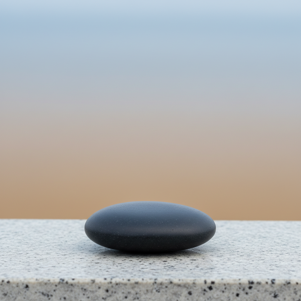 A solitary, perfectly smooth basalt meditation stone, its charcoal surface gently reflecting ambient light, positioned on a pale granite platform. The background is a blurred, subtly graded wash of taupe and misty blue, evoking an airy, zen-inspired sanctuary. Cool, indirect lighting creates soft highlights along the stone’s rounded curves, accentuating its calming presence without harsh contrasts. The image is captured from a slightly elevated angle, employing symmetrical composition for a sense of balance and harmony. The mood is meditative and poised, underscoring holistic tranquility through minimalist design and photographic realism.